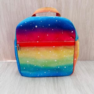 May include: A rainbow-coloured lunch bag with a gradient design, transitioning from blue to red to yellow. It has a red zip and a matching handle. White speckles are scattered across the surface, resembling stars.