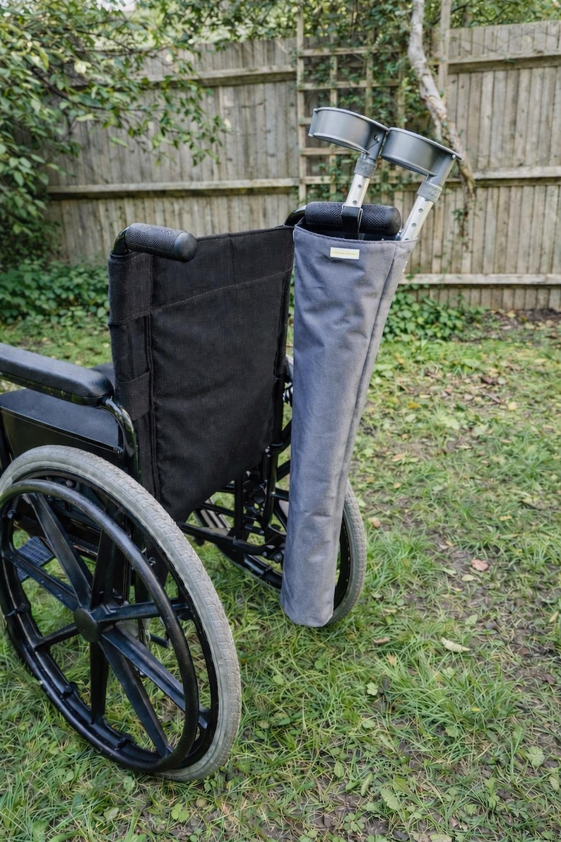 Pu&ograve; includere: Una sedia a rotelle nera con un supporto in tessuto grigio per stampelle fissato sul retro. Il supporto contiene due stampelle antibrachiali in metallo grigio con polsini in plastica nera. La sedia presenta sedile, schienale e ruote nere.