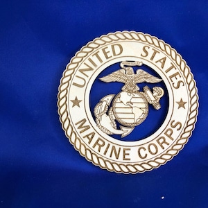 May include: Wooden emblem of the United States Marine Corps, featuring the eagle, globe, and anchor emblem within a circular frame. The words "United States Marine Corps" are engraved around the emblem. The emblem is set against a blue background.