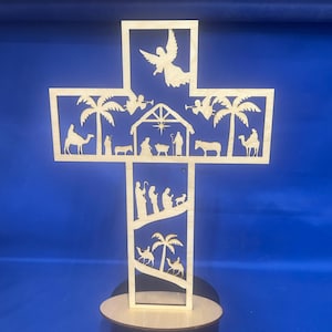 May include: A wooden cross-shaped nativity scene with intricate cut-out designs. The cross features palm trees, angels, and depictions of the nativity story, including the baby Jesus, Mary, Joseph, and the three wise men. It stands on a round wooden base.