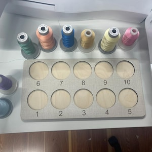 May include: A wooden thread organizer with 10 numbered slots for storing spools of thread. The organizer is gray with white numbers. There are 10 spools of thread in various colors, including pink, blue, green, yellow, and orange.