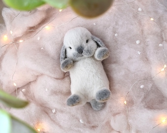 May include: A white and gray felt bunny toy with a sweet expression, lying on a pink fuzzy blanket with white lights.