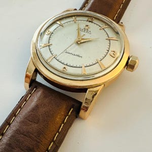 May include: A vintage gold-toned Omega Seamaster automatic wristwatch with a brown leather strap. The watch face is cream-colored with gold hands and markers. The brown leather strap has visible stitching.