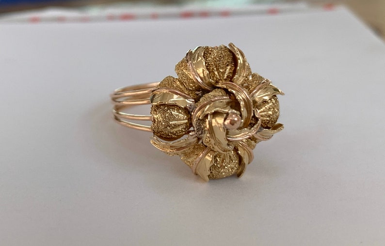 May include: A gold ring with a large, intricate flower design. The flower is made of multiple layers of gold, and the center of the flower has a small, round, gold stone.