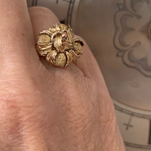 May include: A gold ring with a rose-shaped design. The rose is made of textured gold and has a small red center.