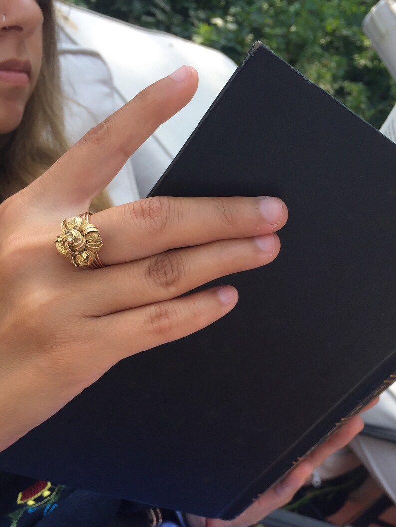 May include: A close-up of a hand wearing a gold ring with a floral design. The hand is holding a black book.