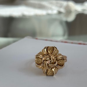 May include: A gold ring with a textured, flower-shaped design. The center of the flower has a small, round stone.