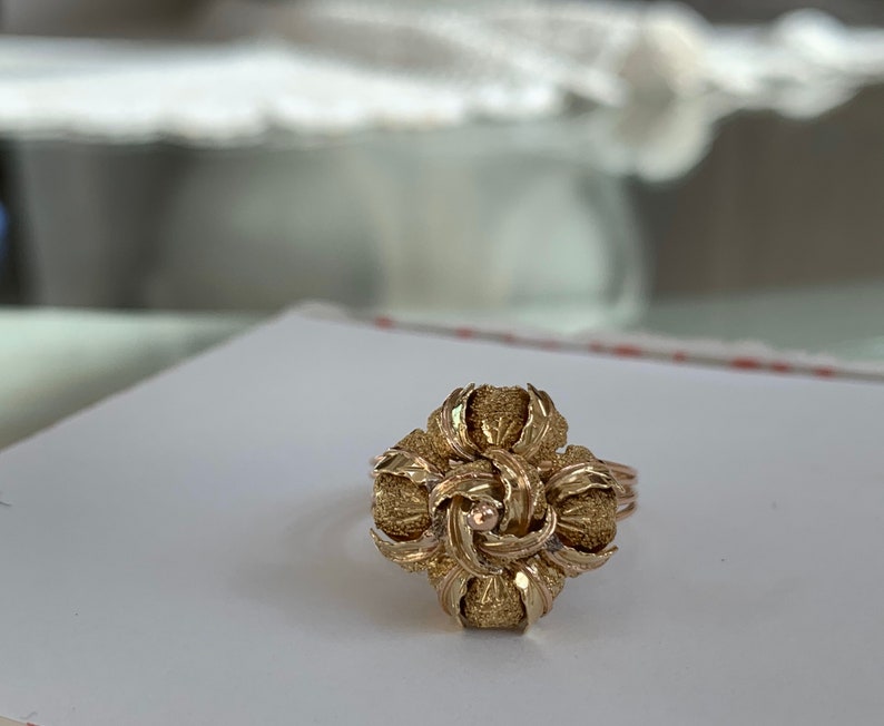 May include: A gold ring with a textured, flower-shaped design. The center of the flower has a small, round stone.