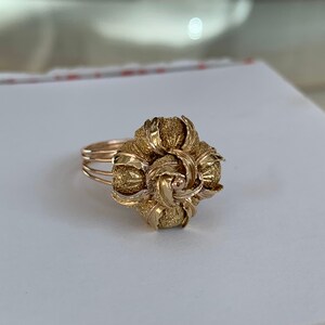 May include: A gold ring with a large, intricate floral design. The ring is made of textured gold and has a delicate band.