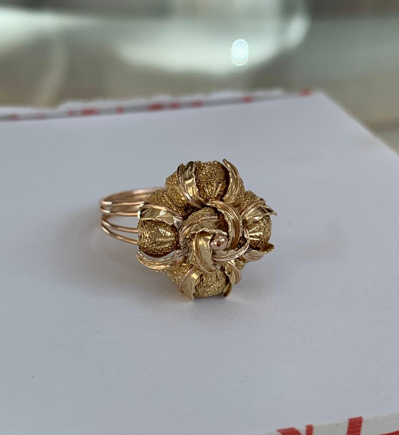 May include: A gold ring with a large, intricate floral design. The ring is made of textured gold and has a delicate band.