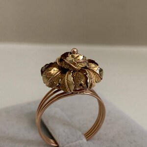May include: A gold ring with a flower-shaped design. The flower is made of multiple layers of leaves, with a textured surface. The ring is made of two-tone gold, with a rose gold band and a yellow gold flower.