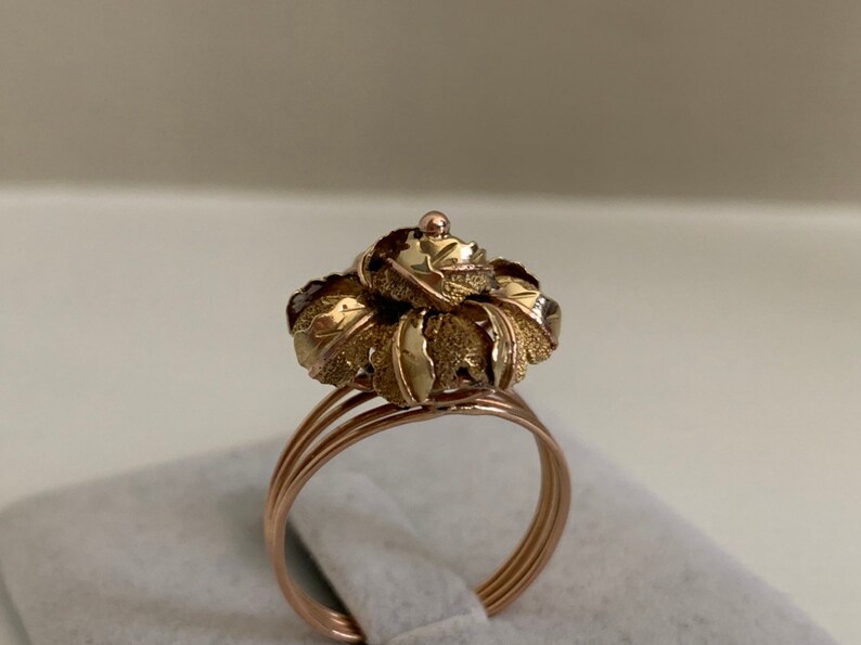 May include: A gold ring with a flower-shaped design. The flower is made of multiple layers of leaves, with a textured surface. The ring is made of two-tone gold, with a rose gold band and a yellow gold flower.