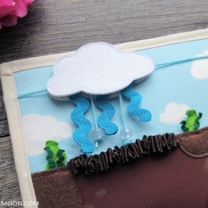 May include: A felt play mat featuring a white cloud and blue raindrops. The mat has a brown felt section with green felt plants and a blue felt sky section. The cloud is attached to the mat with a string and the raindrops are attached to the cloud with strings.
