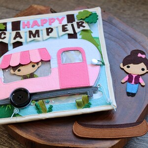 May include: A felt book for children featuring a pink camper van, a girl character, and a canoe. The book has the words "Happy Camper" on the front.