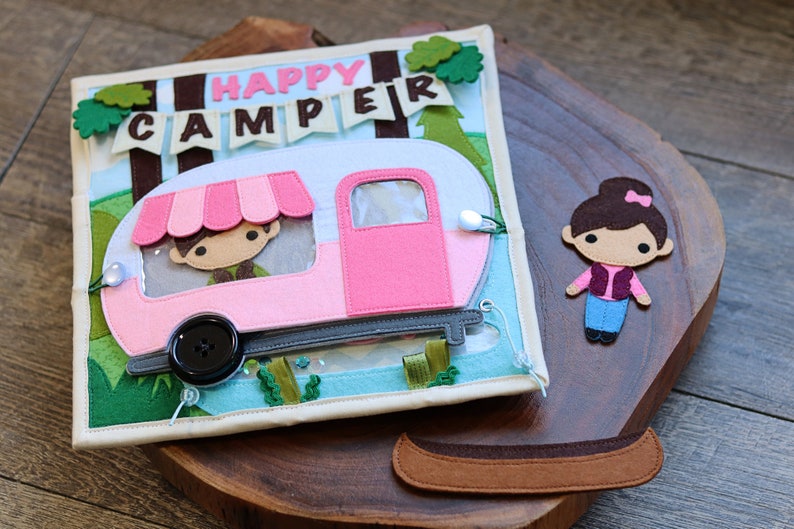 May include: A felt book for children featuring a pink camper van, a girl character, and a canoe. The book has the words "Happy Camper" on the front.