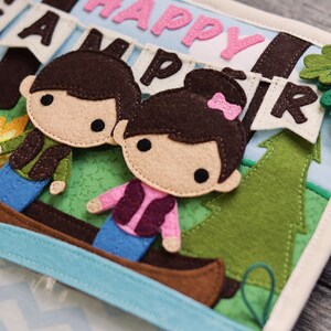 May include: A felt book with a blue and green background. The book features a felt canoe with two felt figures inside. The figures are a boy and a girl with brown hair. The book also features a felt tree and a felt banner that says "Happy Camper".