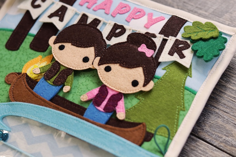 May include: A felt book with a blue and green background. The book features a felt canoe with two felt figures inside. The figures are a boy and a girl with brown hair. The book also features a felt tree and a felt banner that says "Happy Camper".