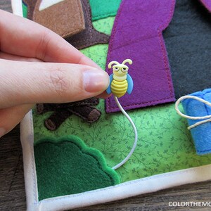 May include: A felt play mat with a purple tent, a blue sleeping bag, a green bush, and a yellow bee with a white string attached to it.