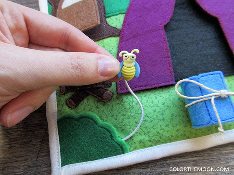 May include: A felt play mat with a purple tent, a blue sleeping bag, a green bush, and a yellow bee with a white string attached to it.