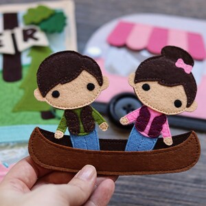 May include: Two felt figures of people in a brown canoe. The figures are wearing blue jeans, a green vest, and a pink vest. The canoe is brown with a blue bottom.