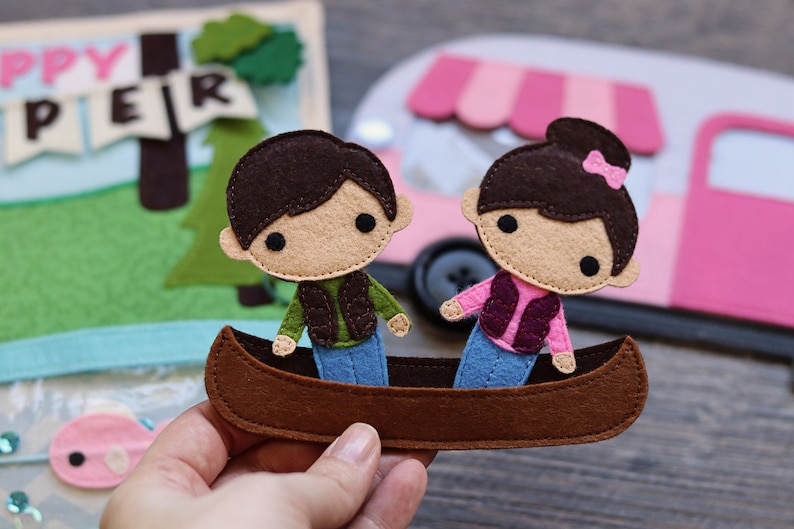 May include: Two felt figures of people in a brown canoe. The figures are wearing blue jeans, a green vest, and a pink vest. The canoe is brown with a blue bottom.