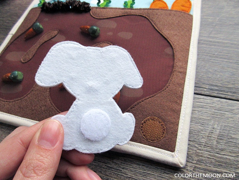 May include: A white felt bunny with a white felt tail, the bunny is attached to a brown felt background with a white felt border.