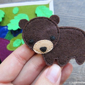 May include: A brown felt bear applique with black eyes and a beige nose. The bear is sitting on a hand.