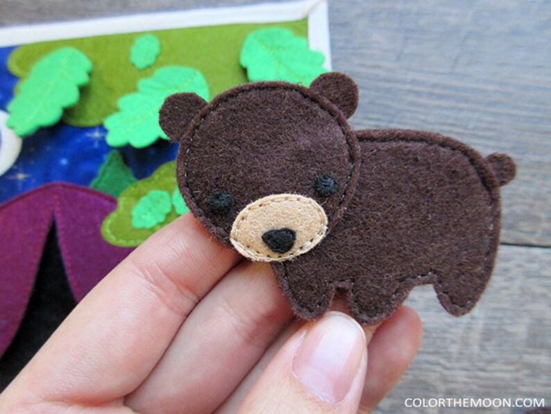 May include: A brown felt bear applique with black eyes and a beige nose. The bear is sitting on a hand.