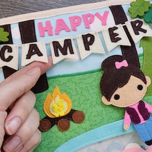 May include: A felt craft depicting a camping scene with a girl, a campfire, and a banner that reads "Happy Camper".