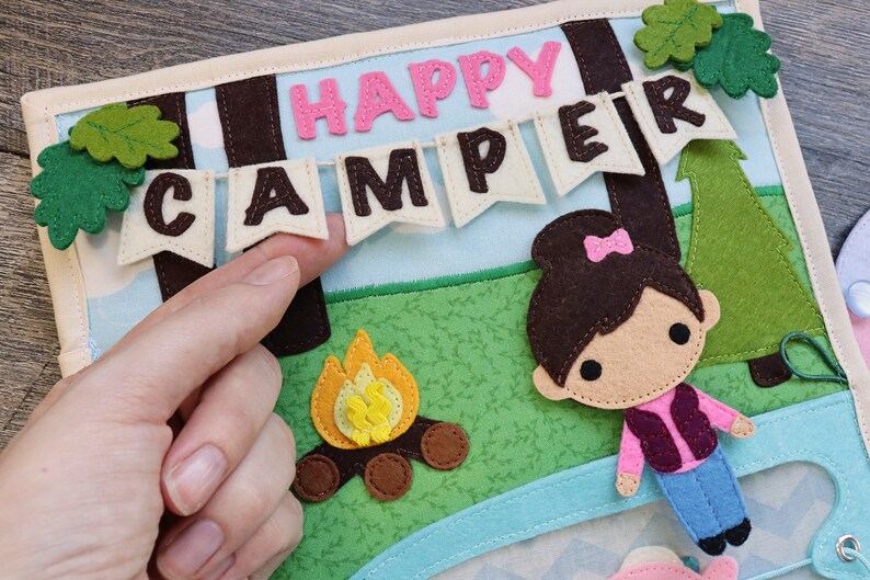 May include: A felt craft depicting a camping scene with a girl, a campfire, and a banner that reads "Happy Camper".