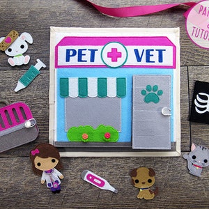 May include: Felt play set for pretend play. The set features a pink and blue pet vet clinic with a green awning, a veterinarian, a pet carrier, a syringe, a thermometer, a dog, a cat, and an x-ray.