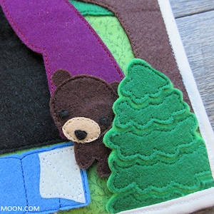 May include: A felt appliqu&eacute; of a brown bear sleeping in a blue sleeping bag with a green felt tree behind it. The bear has a brown nose and black eyes. The sleeping bag has a white flap. The tree has a green felt texture.