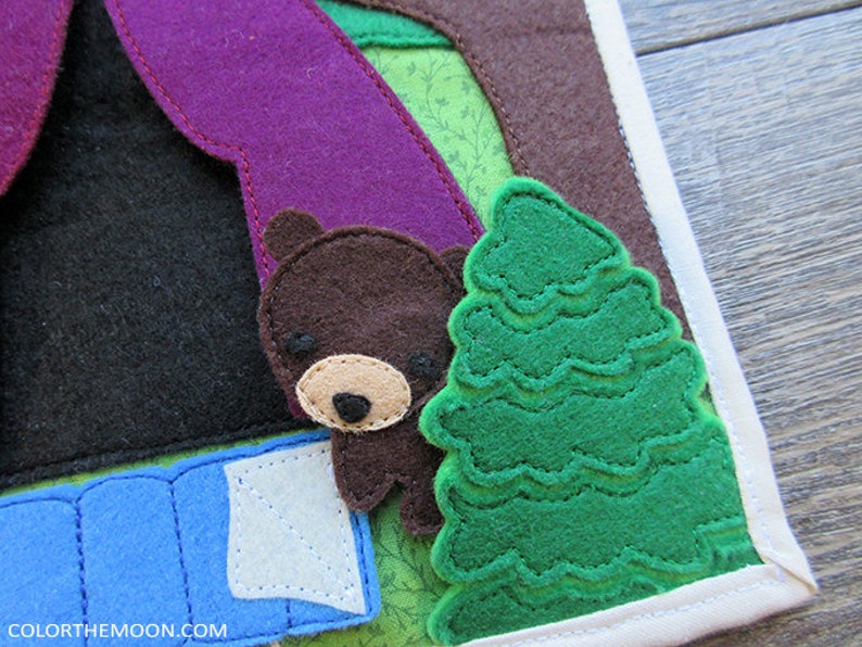 May include: A felt appliqu&eacute; of a brown bear sleeping in a blue sleeping bag with a green felt tree behind it. The bear has a brown nose and black eyes. The sleeping bag has a white flap. The tree has a green felt texture.