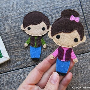 May include: Two felt figures of children, a boy and a girl, wearing brown vests and blue jeans. The girl has a pink bow in her hair.