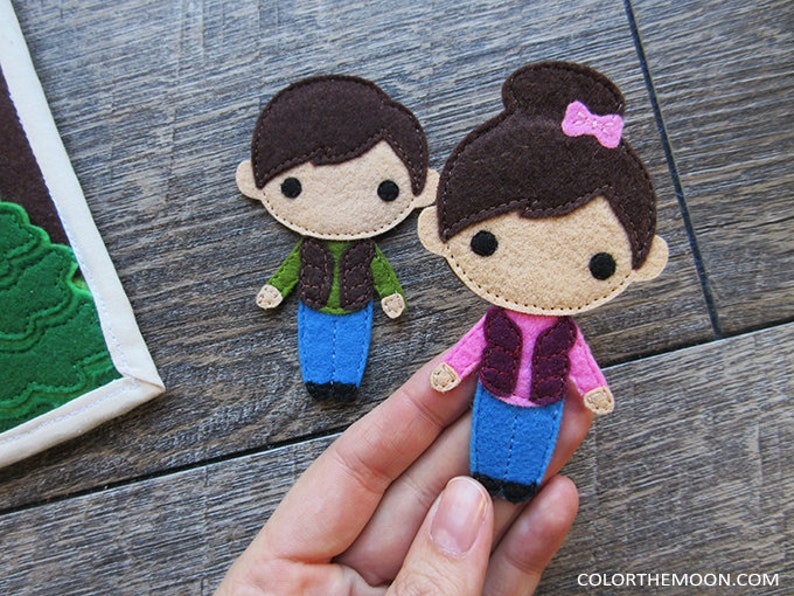 May include: Two felt figures of children, a boy and a girl, wearing brown vests and blue jeans. The girl has a pink bow in her hair.