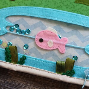 May include: A blue felt board with a pink felt fish, green felt seaweed, and blue sequins. The fish is attached to a string that is threaded through two metal eyelets.