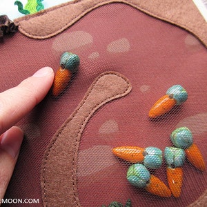 May include: A close-up of a brown felt board with a mesh overlay. The board is decorated with orange felt carrots and turquoise felt beads. The board is designed for children to play with and learn about colours and shapes.