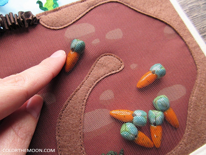 May include: A close-up of a brown felt board with a mesh overlay. The board is decorated with orange felt carrots and turquoise felt beads. The board is designed for children to play with and learn about colours and shapes.
