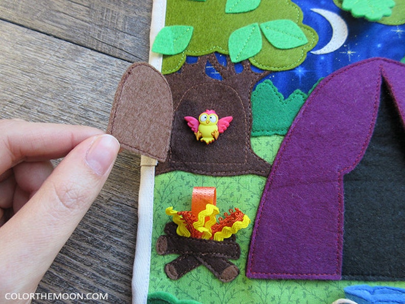 May include: A felt play mat with a purple tent, a brown tree with a birdhouse, a campfire, and green leaves. The tree has a small, pink bird with yellow accents sitting in the birdhouse.