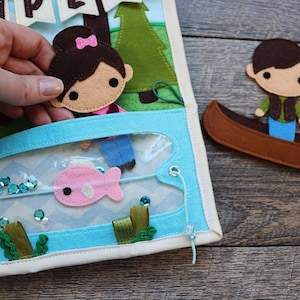 May include: A felt play set featuring a girl and a boy character. The girl is holding a fishing rod and there is a pink fish in the water. The boy is in a canoe. The set is designed for imaginative play.