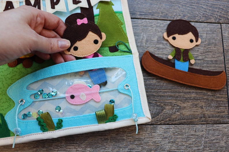 May include: A felt play set featuring a girl and a boy character. The girl is holding a fishing rod and there is a pink fish in the water. The boy is in a canoe. The set is designed for imaginative play.