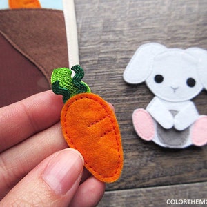 May include: A felt carrot with green leaves, a felt bunny with pink feet, and a brown felt patch of dirt. The carrot is orange and has a stitched-on design.