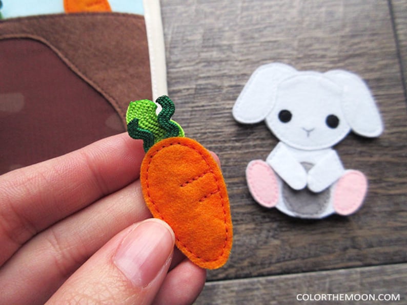 May include: A felt carrot with green leaves, a felt bunny with pink feet, and a brown felt patch of dirt. The carrot is orange and has a stitched-on design.