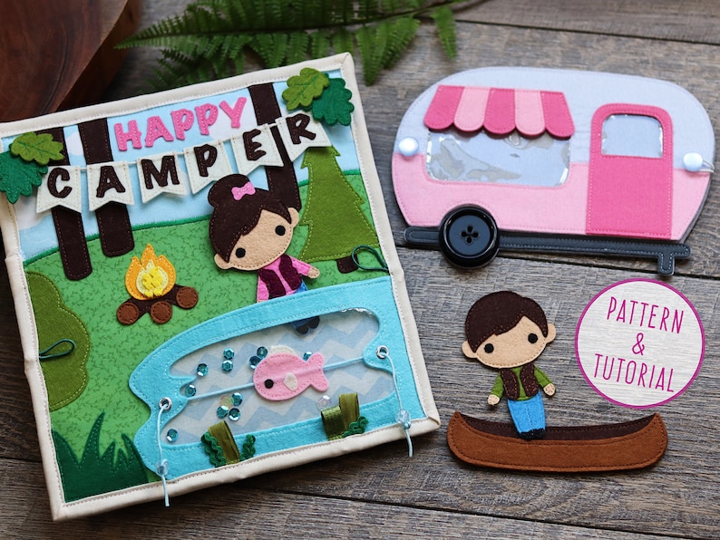 May include: A felt book with a camping theme. The book features a girl with brown hair and a pink bow, a campfire, a lake with a fish, and a camper. The book also includes a separate felt camper and a felt boy in a canoe.