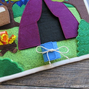 May include: A felt play mat with a purple tent, a campfire, a green tree, and a blue roll of fabric tied with twine. The mat is surrounded by a white border.