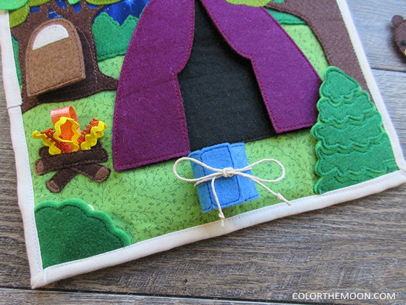 May include: A felt play mat with a purple tent, a campfire, a green tree, and a blue roll of fabric tied with twine. The mat is surrounded by a white border.