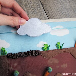 May include: A felt play set with a blue sky, white clouds, brown dirt, green grass, and orange carrots. The cloud is attached to the sky with a blue string and can be moved around.