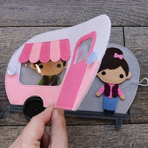May include: A pink and white felt camper with a pink awning and a clear window. The camper has a pink door that is open. A felt figure of a person is inside the camper. A felt figure of a person is standing outside the camper.
