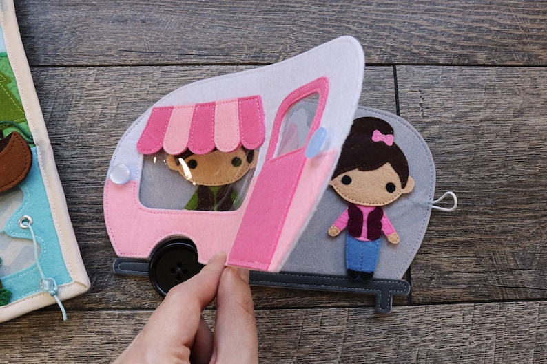 May include: A pink and white felt camper with a pink awning and a clear window. The camper has a pink door that is open. A felt figure of a person is inside the camper. A felt figure of a person is standing outside the camper.