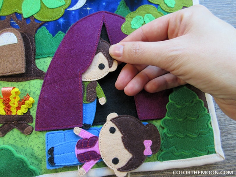 May include: A felt play set with a purple tent, a campfire, and two figures. The figures are a boy and a girl with brown hair. The boy is wearing a brown jacket and blue jeans. The girl is wearing a pink shirt and blue jeans. The tent is open and the boy is peeking out.